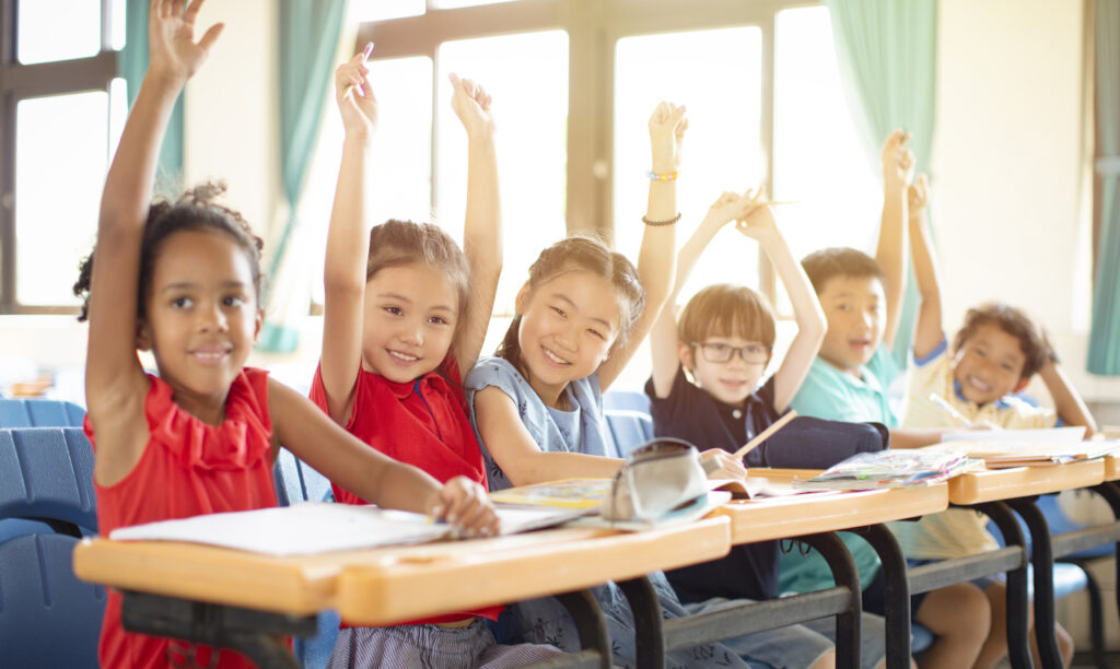 Young elementary school-aged children raising their hands in a classroom